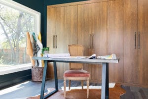 Well lit office with modern desk and antique chair staged with an animal skin rug in front of a warm natural would Murphy bed folded away.