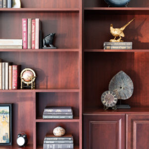 Cherrywood bookshelves staged with books stack vertically and horizontally featuring unique bookends: a globe, an alarm clock, a vase, black frog, gold bird, and geodes.