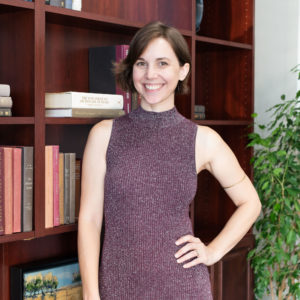 More Space Place designer Cyndi Nelvin in a mauve knit mock neck dress posed in front of cherrywood bookshelves and a plant.